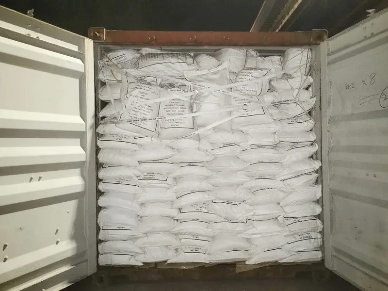 Open shipping container stacked to the roof with white 30 kg bags held in place by lashing straps